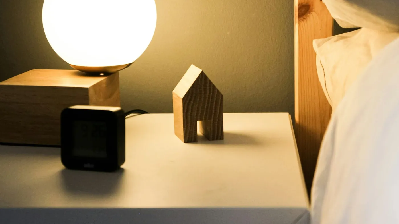 Una lámpara encendida y un reloj despertador sobre una mesa de luz, junto a una cama
