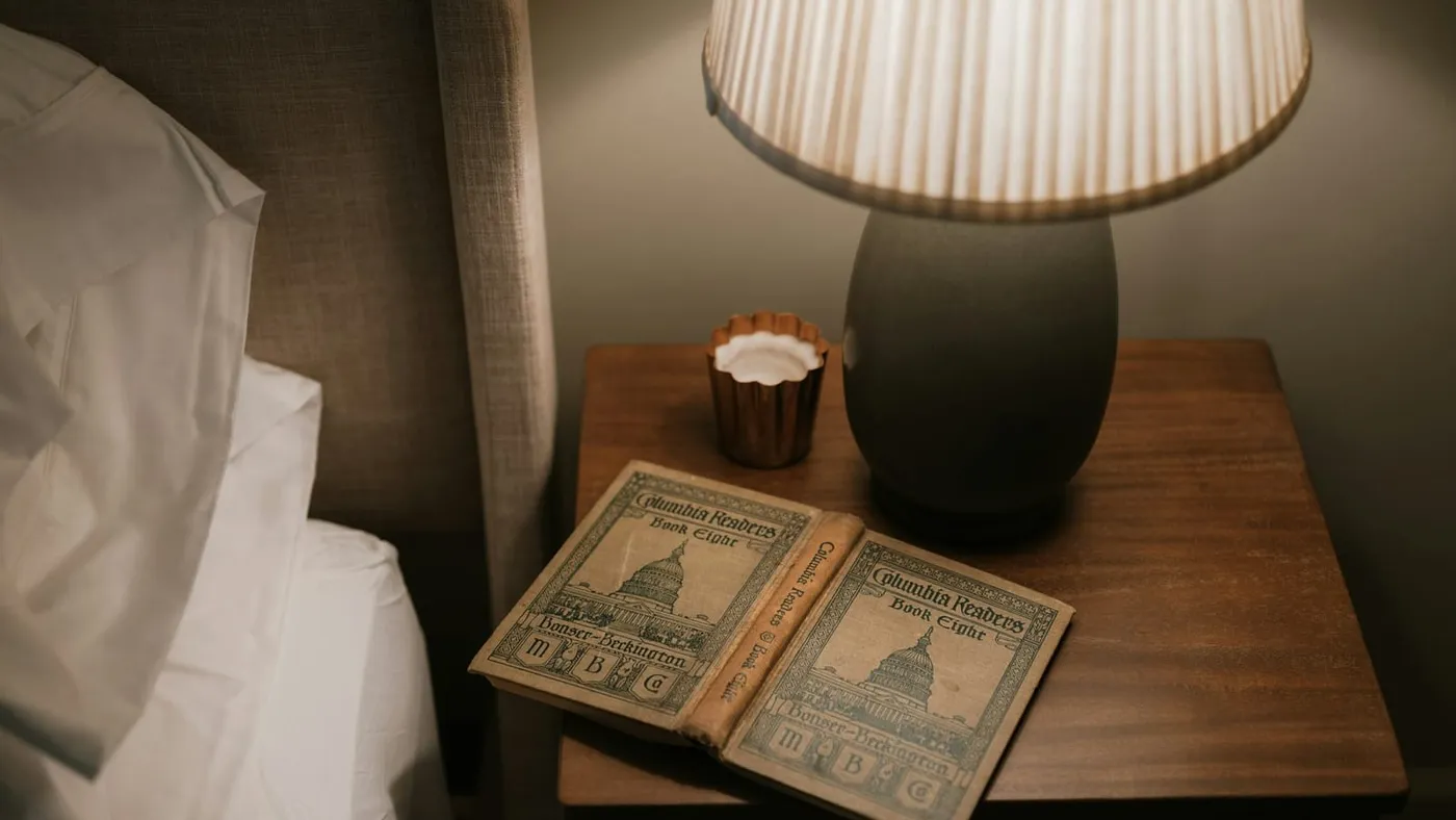 Una mesa de luz con un velador encendido y un libro, junto a una cama