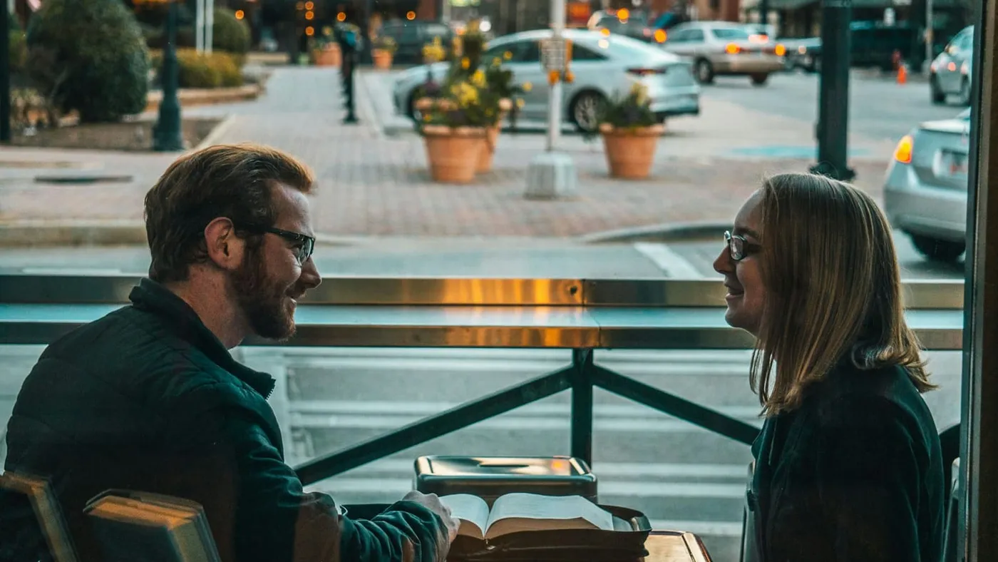 Un hombre y una mujer conversan sentados frente a frente junto a la ventana de un café