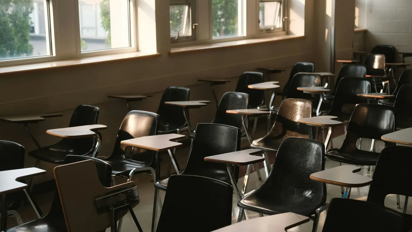 Un aula vacía con pupitres y sillas junto a una fila de ventanas por las que entra luz