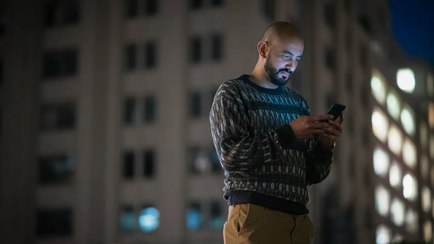 Un hombre joven mira su celular de noche, con edificios iluminados de fondo