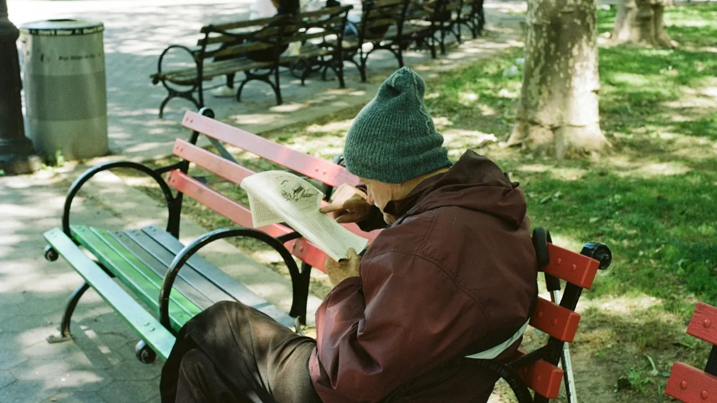 Una persona con gorro de lana y campera lee sentada en un banco de plaza