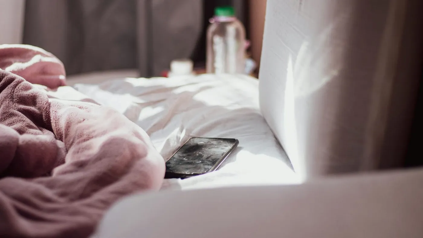 Un celular apoyado sobre una cama deshecha, junto a una frazada