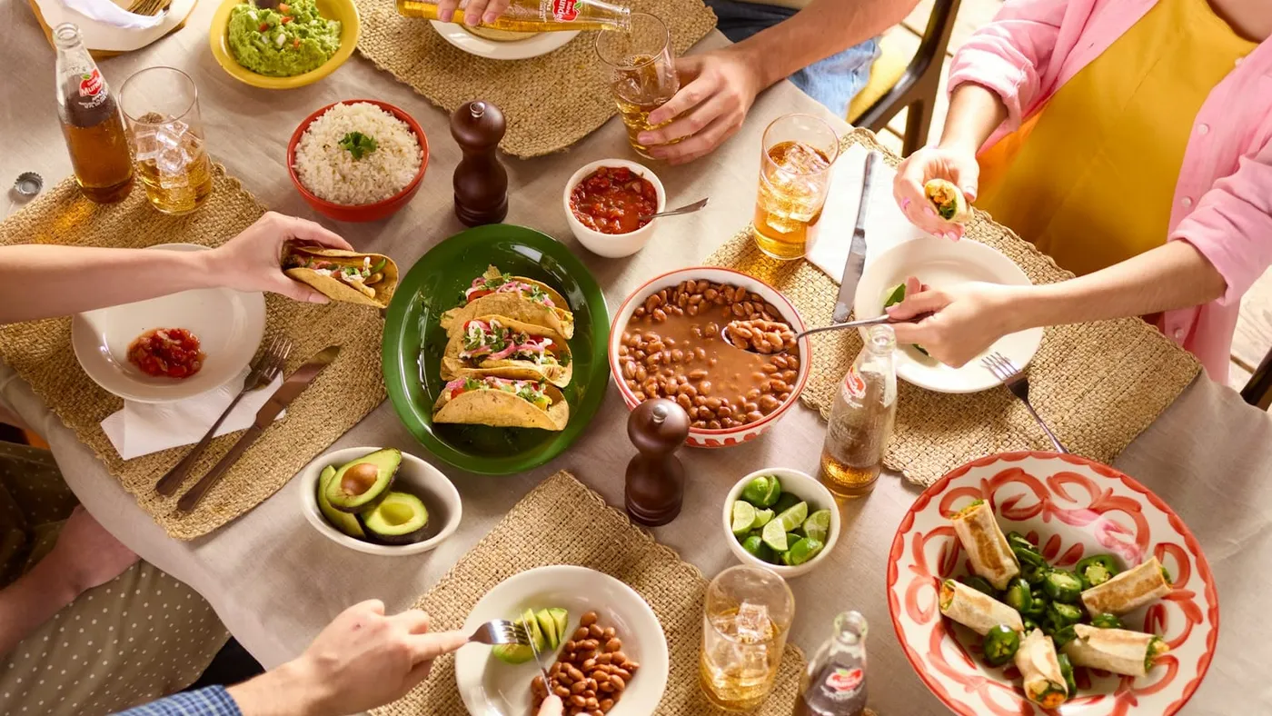 Vista desde arriba de una mesa compartida, con varias manos sirviéndose de platos y fuentes de comida