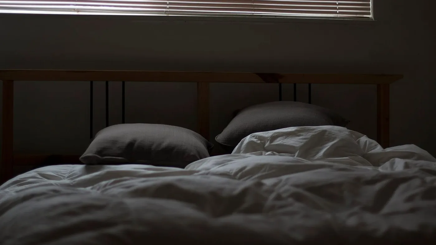 Una cama deshecha en un cuarto en penumbra, con la persiana baja